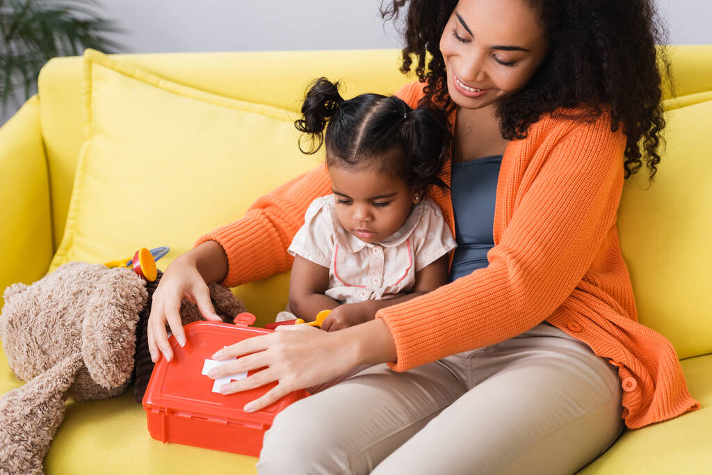 happy african american mother sitting with toddler daughter and holding toy first aid kit in living room - Photo, Image