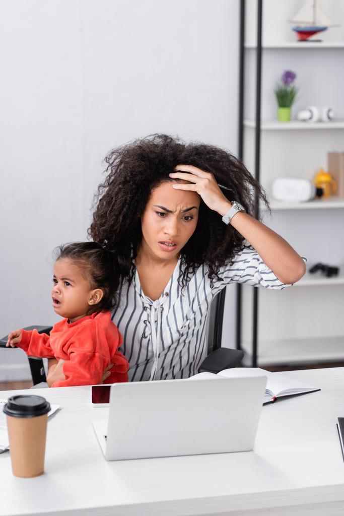 stressed african american freelancer holding in arms crying toddler daughter while working from home  - Photo, Image