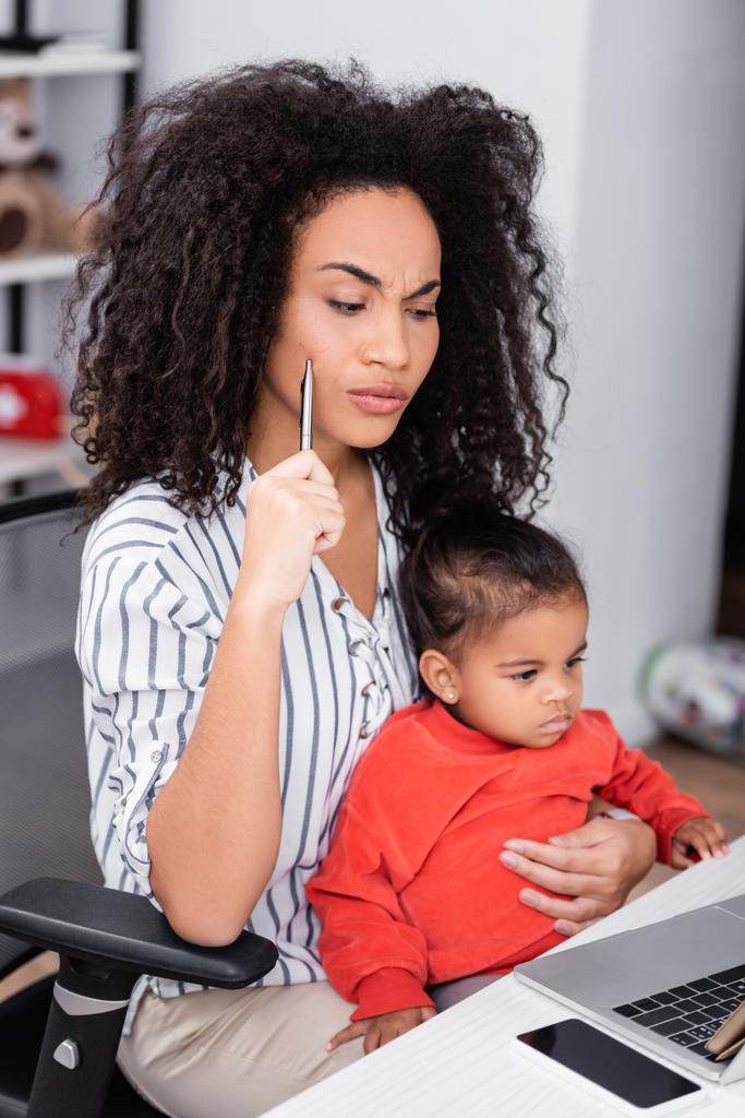 σκεπτική αφρικάνικη αμερικανική μητέρα κρατώντας στυλό και κάθεται με την κόρη νήπιο, ενώ εργάζονται από το σπίτι  - Φωτογραφία, εικόνα