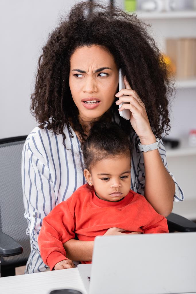 Afrykańska amerykańska matka rozmawia na smartfonie siedząc z córką malucha w pobliżu laptopa na niewyraźnym pierwszym planie - Zdjęcie, obraz