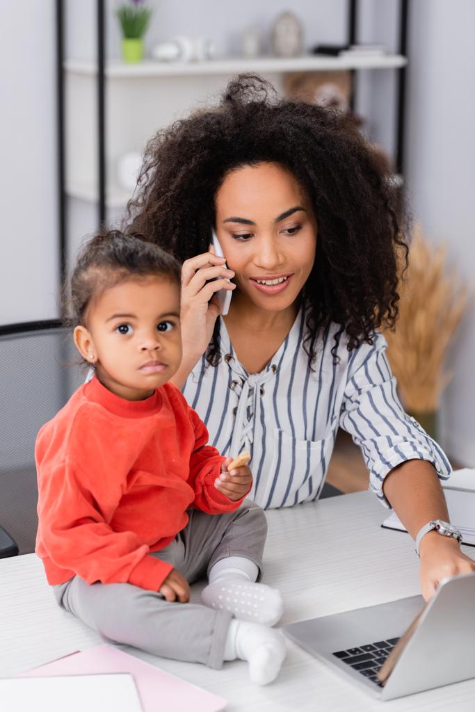 tout-petit enfant afro-américain tenant un cookie savoureux près de la mère parlant sur smartphone tout en travaillant à la maison - Photo, image