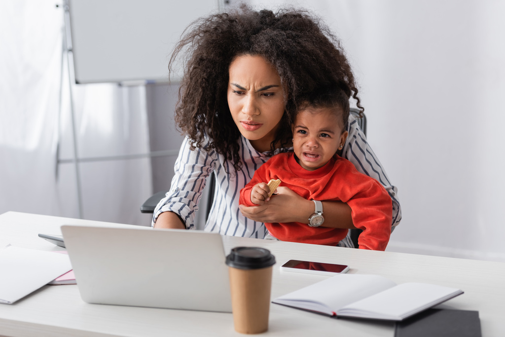 estressado afro-americano mãe segurando em braços chorando criança enquanto trabalhava em casa  - Foto, Imagem
