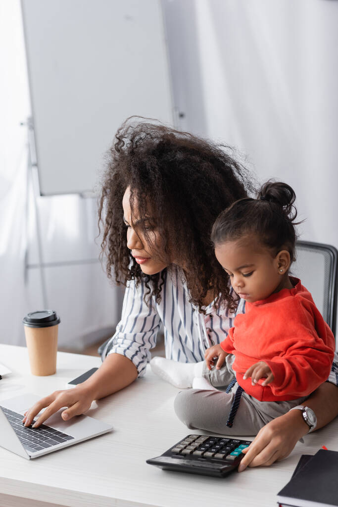 fókuszált afro-amerikai anya a laptop kisgyermek lánya az asztalon - Fotó, kép
