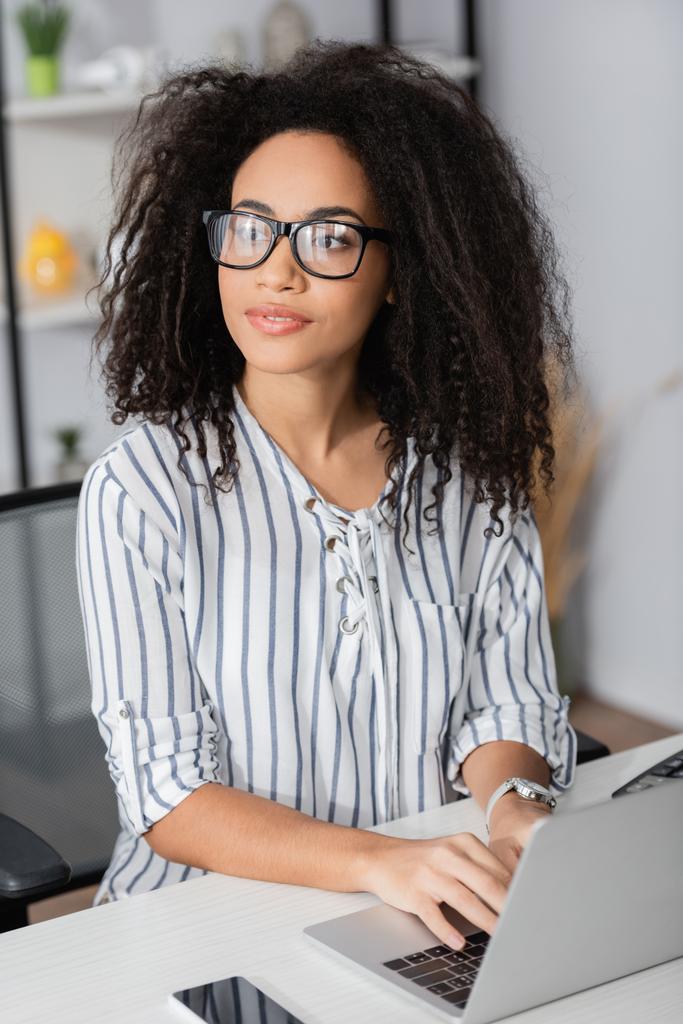freelance afroamericano in occhiali guardando altrove e utilizzando il computer portatile a casa - Foto, immagini