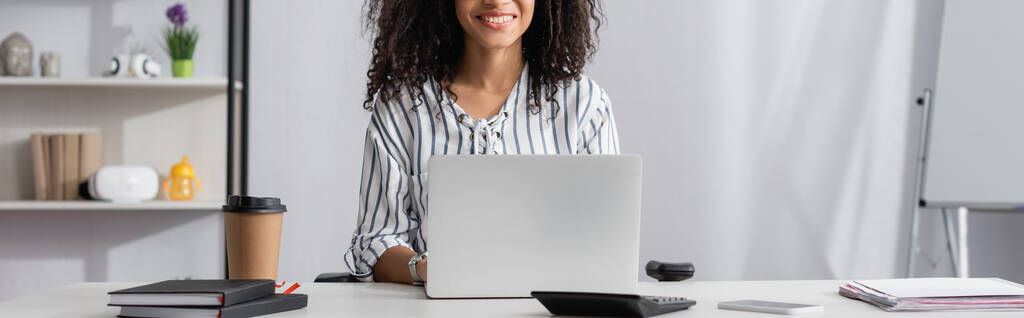 částečný pohled na šťastný africký americký freelancer pomocí notebooku v blízkosti smartphone na stole, banner - Fotografie, Obrázek