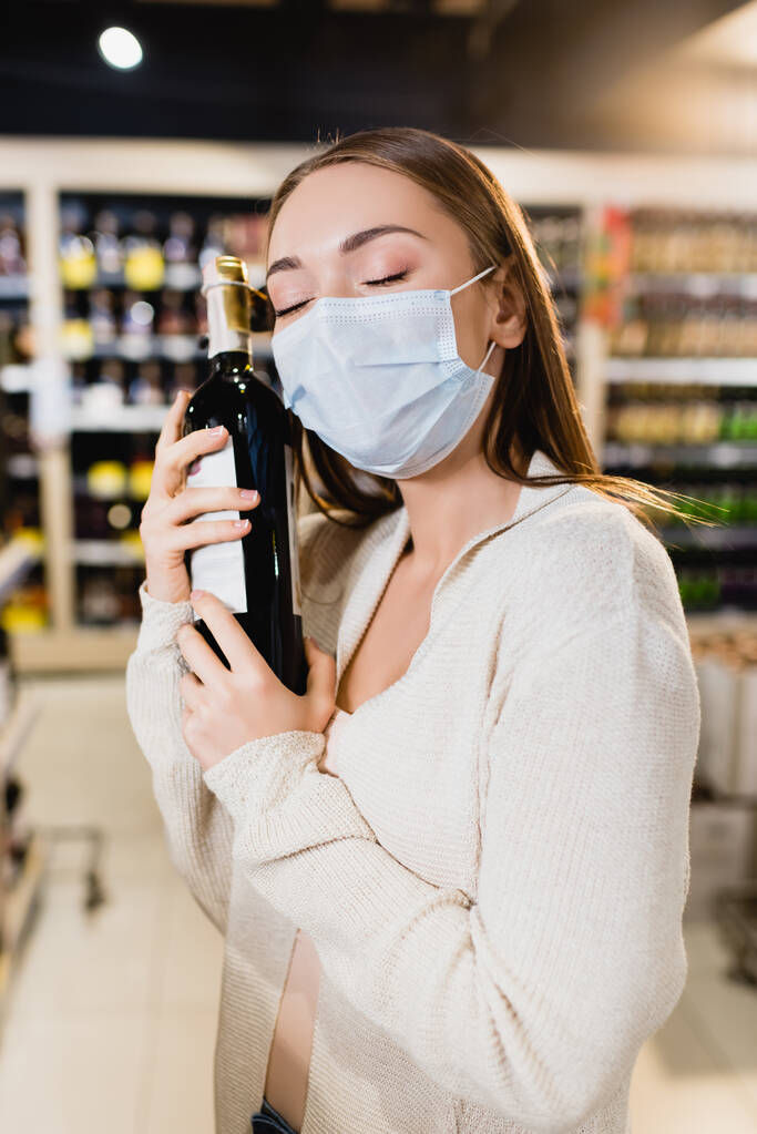 Γυναίκα με ιατρική μάσκα αγκαλιάζει μπουκάλι κρασί στο σούπερ μάρκετ  - Φωτογραφία, εικόνα