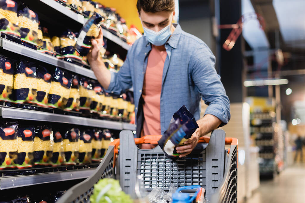 Muž v lékařské masce s potravinami v blízkosti polic a nákupního košíku v supermarketu  - Fotografie, Obrázek