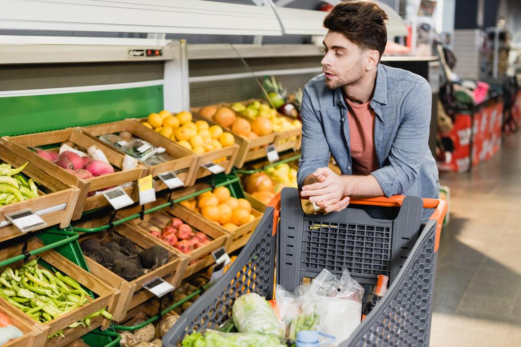 Giovane uomo in piedi vicino al carrello della spesa e frutta fresca sul bancone in negozio  - Foto, immagini