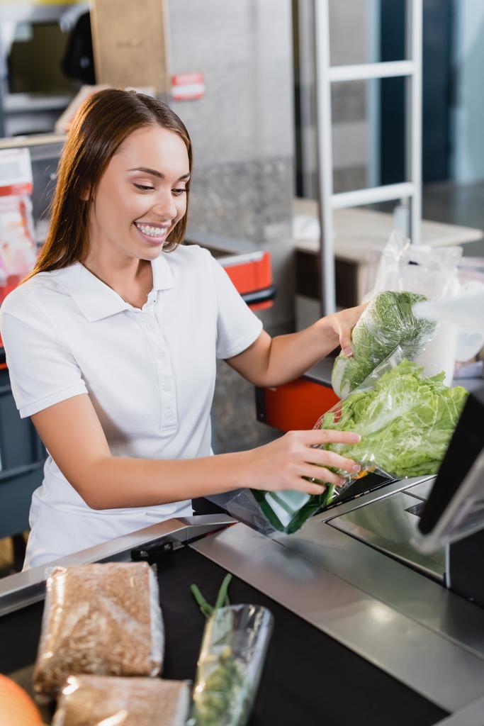 Uśmiechnięty kasjer trzymający warzywa w kasie supermarketu - Zdjęcie, obraz