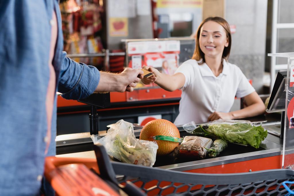 Hombre dando tarjeta de crédito a cajero sonriente cerca de la comida en la caja del supermercado  - Foto, Imagen