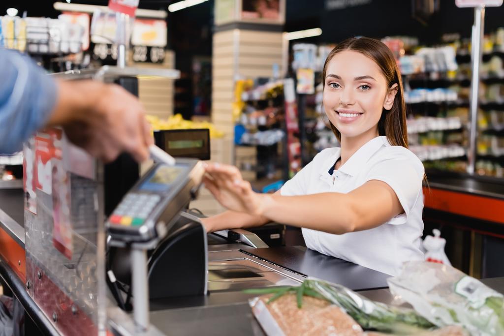 Cajero sonriente mirando la cámara cerca del cliente pagando con teléfono inteligente en primer plano borroso en el supermercado  - Foto, imagen