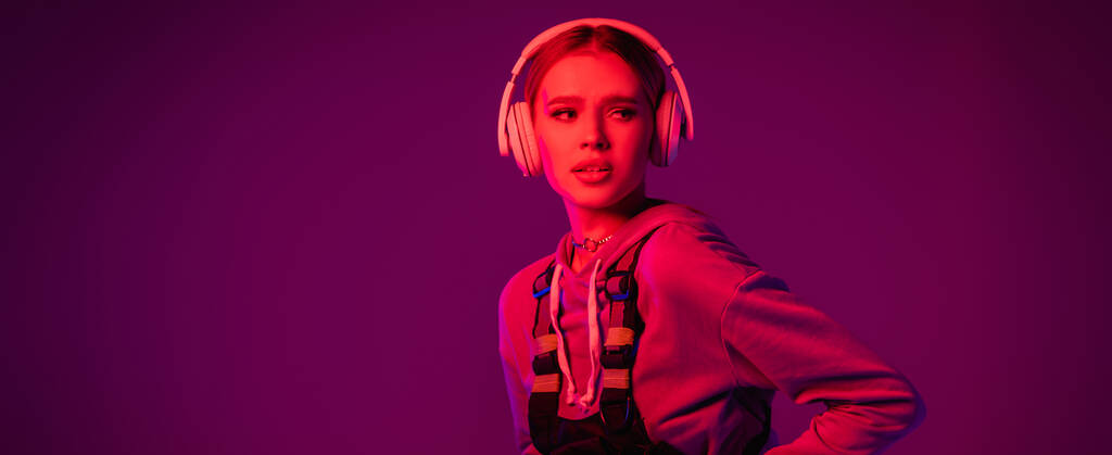 mujer joven en auriculares inalámbricos mirando hacia otro lado sobre fondo púrpura, bandera - Foto, imagen
