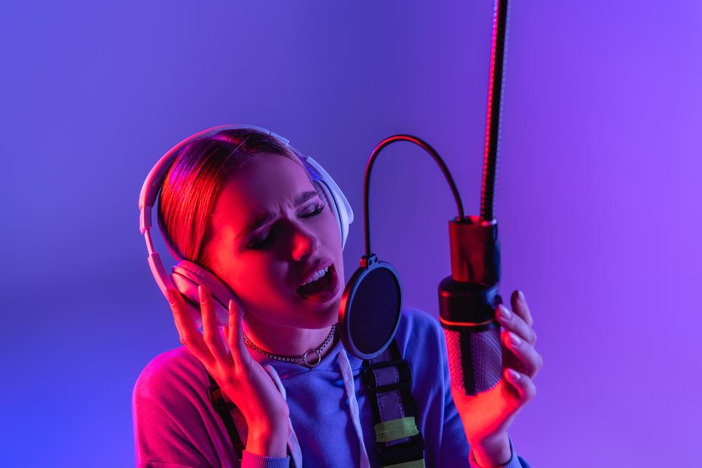 Kulaklıklı genç şarkıcı mikrofonla şarkı söylerken renk filtresiyle mor üzerine şarkı söylüyor.  - Fotoğraf, Görsel