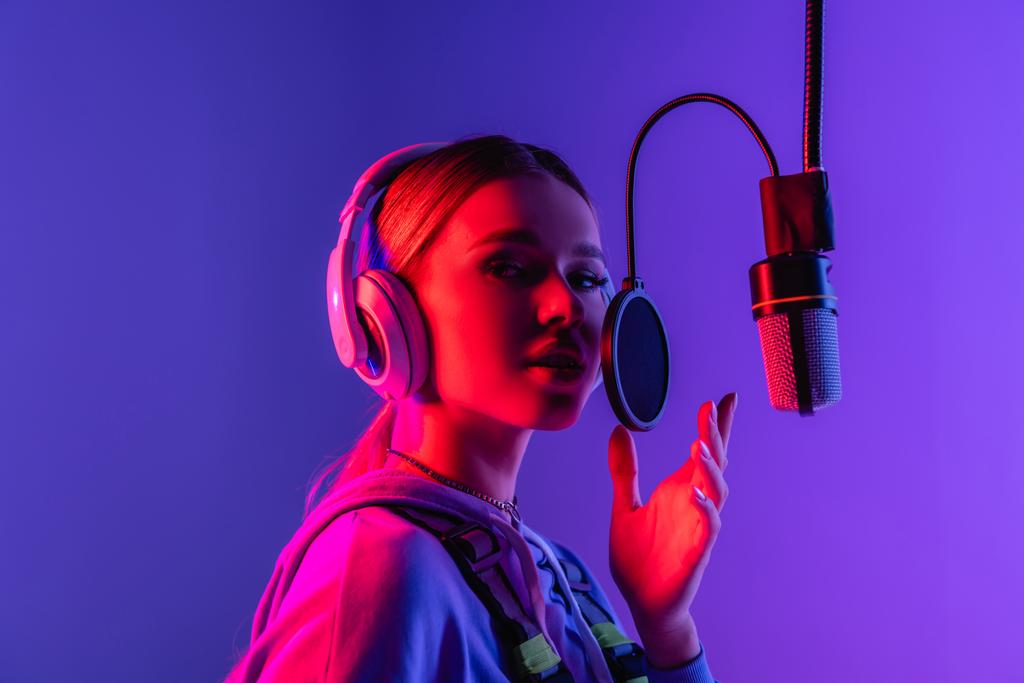 junge Sängerin mit drahtlosen Kopfhörern singt Lied in Mikrofon auf lila  - Foto, Bild