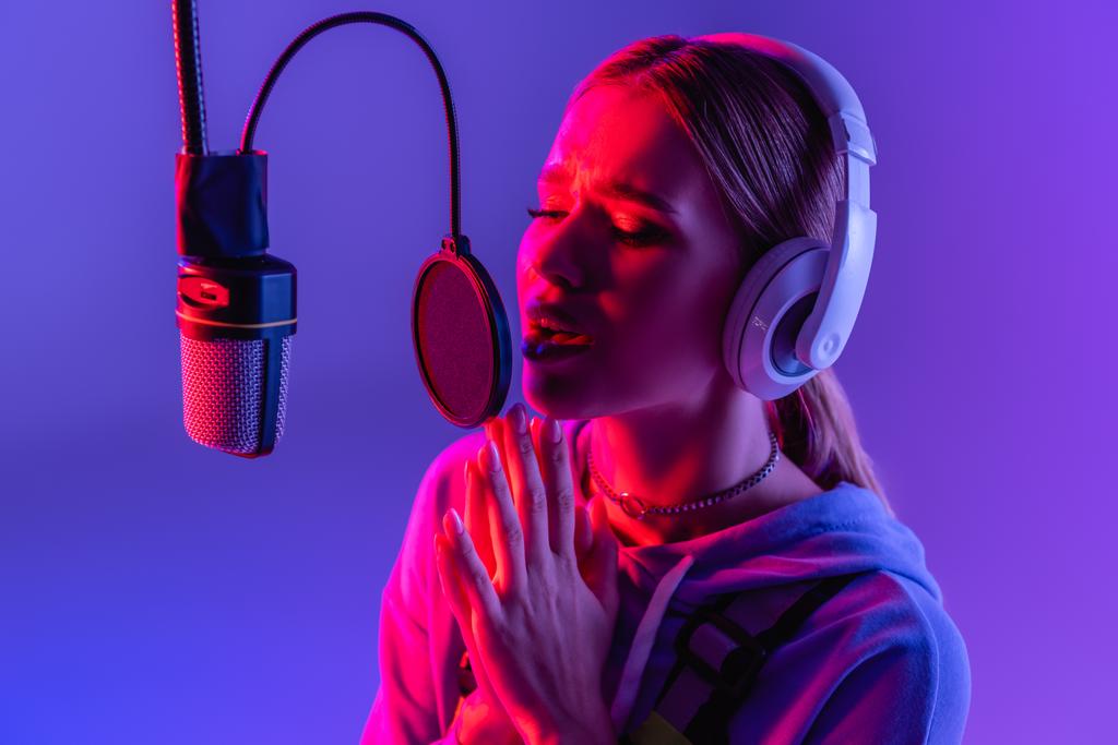 νεαρή γυναίκα σε ασύρματα ακουστικά τραγούδι εγγραφής, ενώ τραγουδούν σε μωβ με φίλτρο χρώματος  - Φωτογραφία, εικόνα