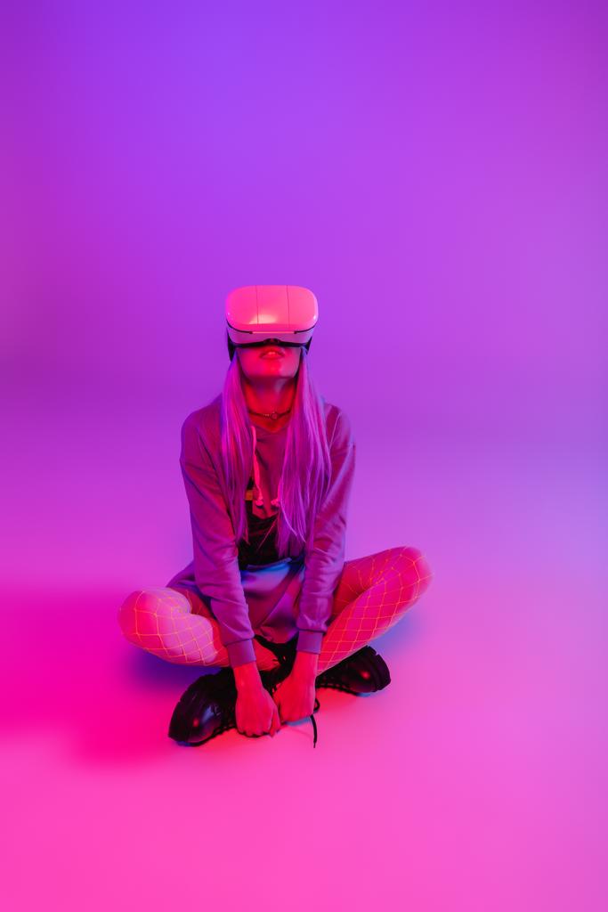 młoda kobieta w wirtualnej rzeczywistości zestaw słuchawkowy siedzi ze skrzyżowanymi nogami na fioletowy i różowy - Zdjęcie, obraz
