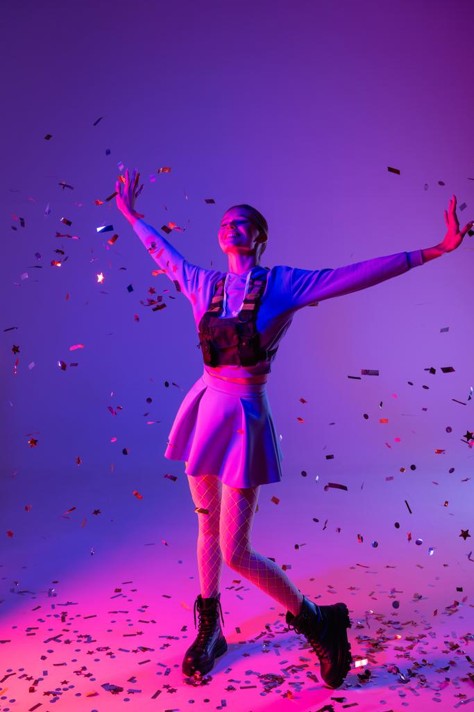 полная длина счастливой женщины в стильном наряде с протянутыми руками рядом падения конфетти на фиолетовый - Фото, изображение