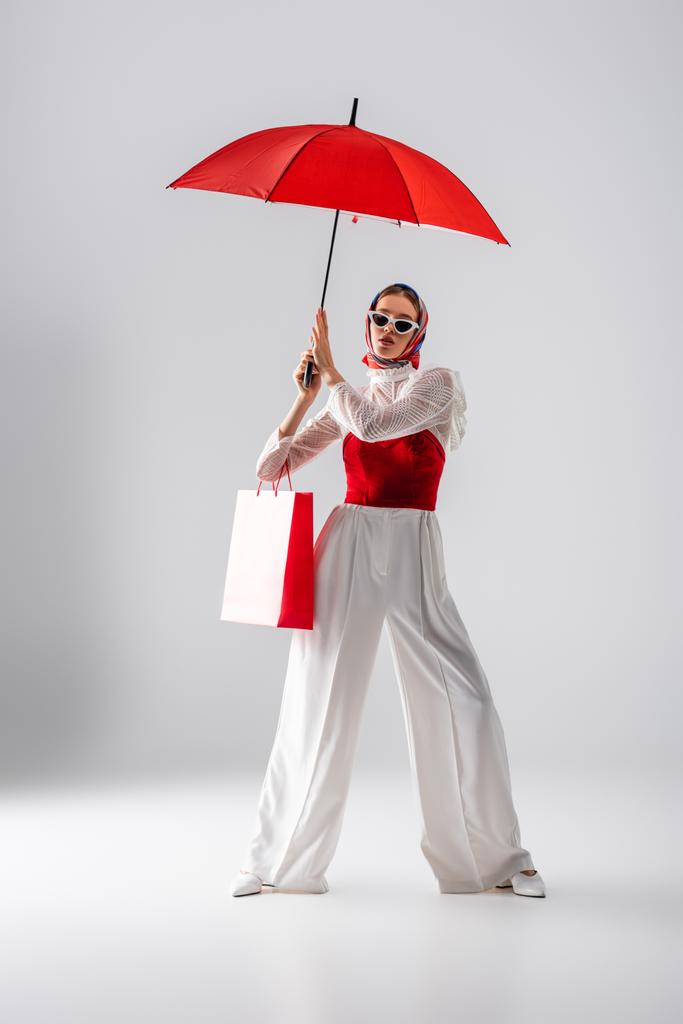 πλήρες μήκος της κομψής γυναίκας σε μαντίλα και γυαλιά ηλίου κρατώντας τσάντα ψώνια, ενώ στέκεται κάτω από ομπρέλα σε λευκό  - Φωτογραφία, εικόνα
