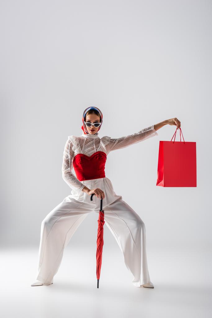 longitud completa de la mujer en pañuelo para la cabeza y gafas de sol de moda sosteniendo paraguas rojo y bolsa de compras mientras posando en blanco  - Foto, imagen