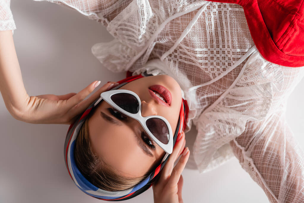top view of trendy woman in sunglasses and headscarf lying while looking at camera on white - Photo, Image