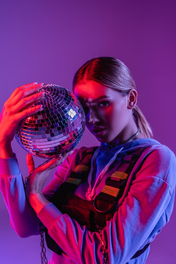 jovem e elegante mulher segurando bola de discoteca brilhante e olhando para a câmera isolada em roxo - Foto, Imagem