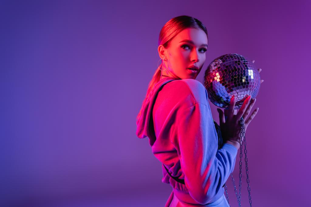 jovem e elegante mulher segurando bola de discoteca brilhante e olhando para longe em roxo - Foto, Imagem