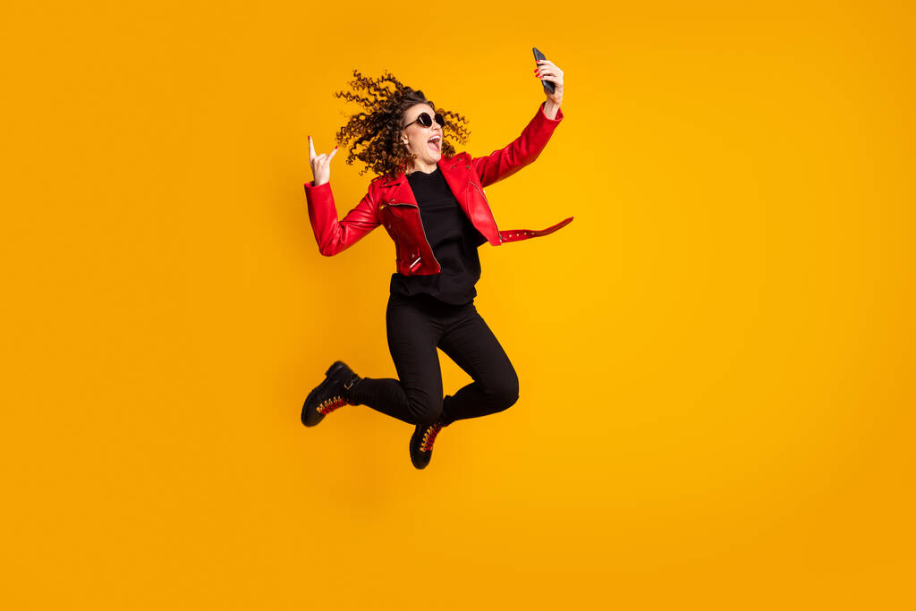 Full length body size view of cheerful wavy-haired girl jumping using cell showing horn sign taking selfie isolated on yellow color background - Photo, Image