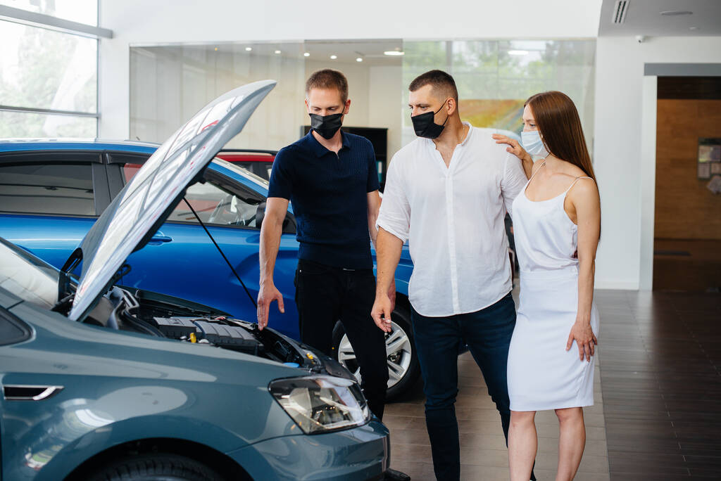 Νεαρό ζευγάρι με μάσκες επιλέγει ένα νέο όχημα και συμβουλεύεται έναν εκπρόσωπο της αντιπροσωπείας κατά την περίοδο της πανδημίας. Πωλήσεις αυτοκινήτων, και τη ζωή κατά τη διάρκεια της πανδημίας - Φωτογραφία, εικόνα