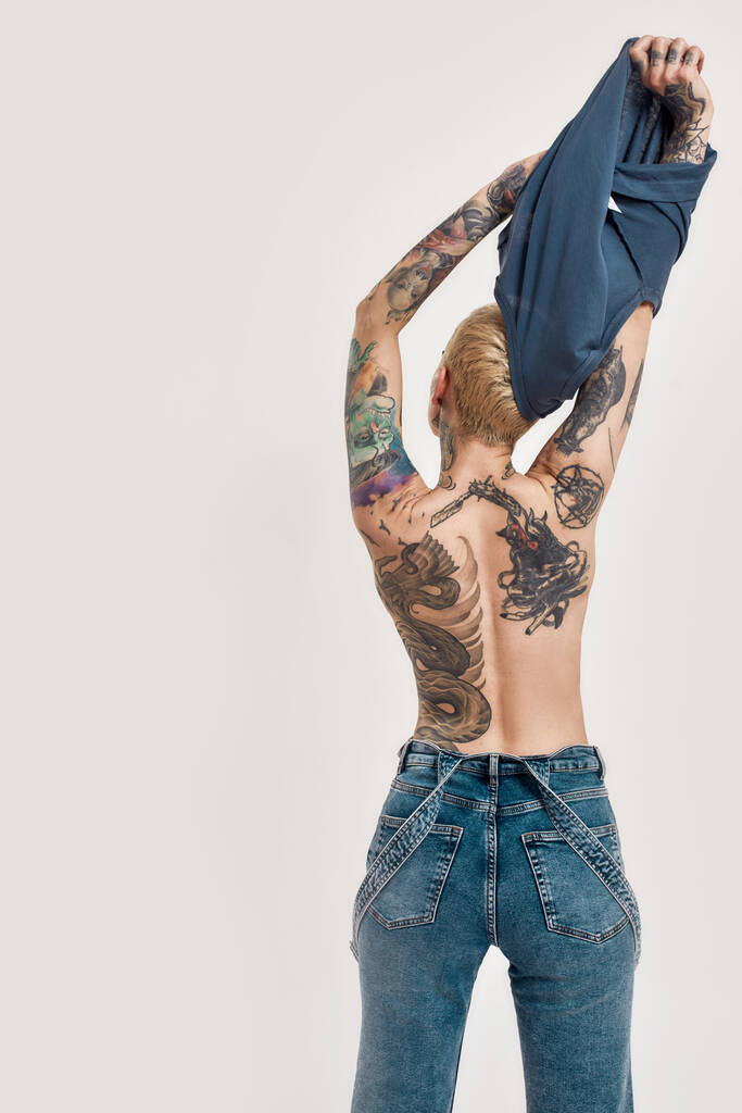 Tatuaż i kolczyk. Przekłuta i wytatuowana biała dziewczyna w dżinsach stojąca tyłem do kamery i kończąca zdjęcia niebieskiej koszulki - Zdjęcie, obraz