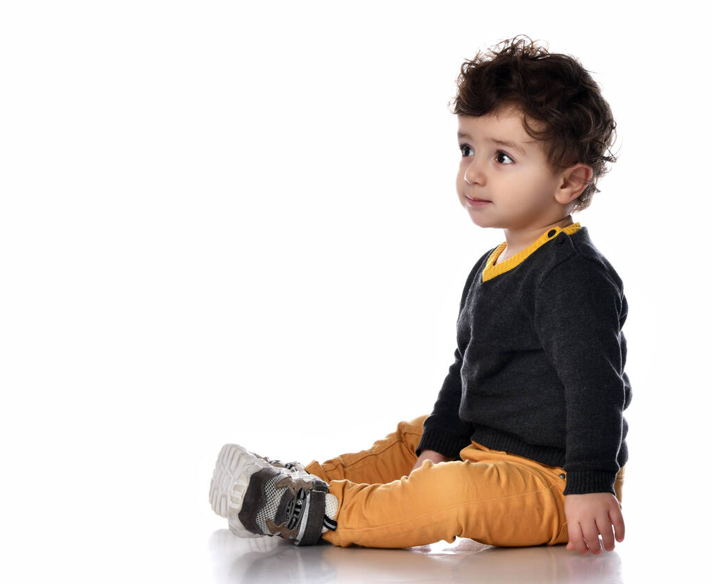 Niño en pantalones amarillos se sienta de costado en el suelo mirándonos contra un fondo blanco con espacio para texto libre - Foto, Imagen