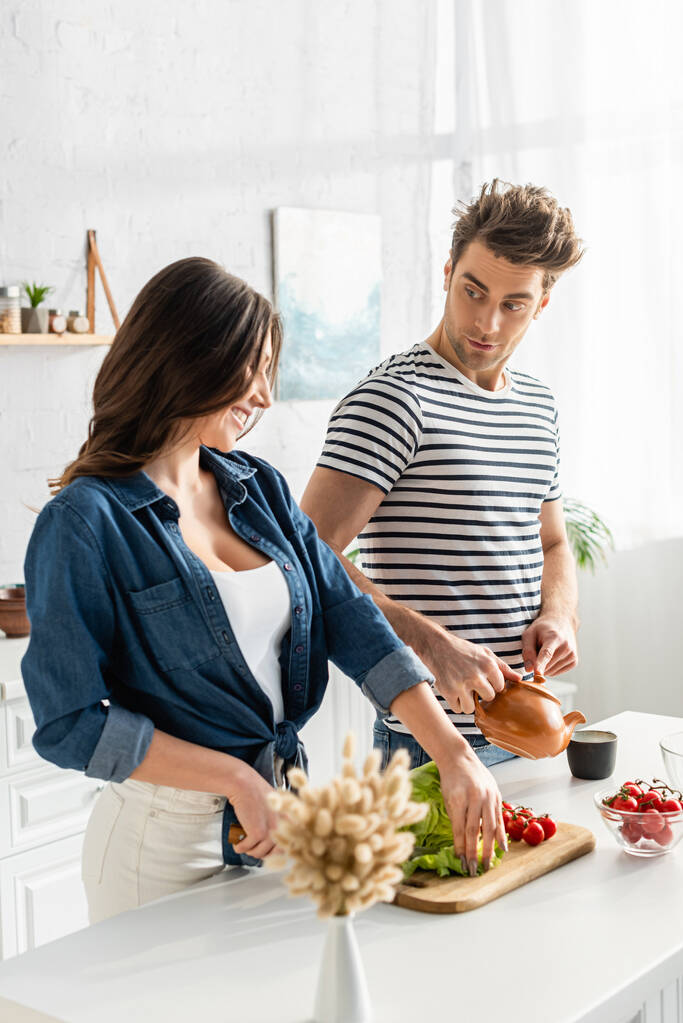 счастливая женщина режет салат рядом с ингредиентами на кухонном столе и парень готовит чай - Фото, изображение