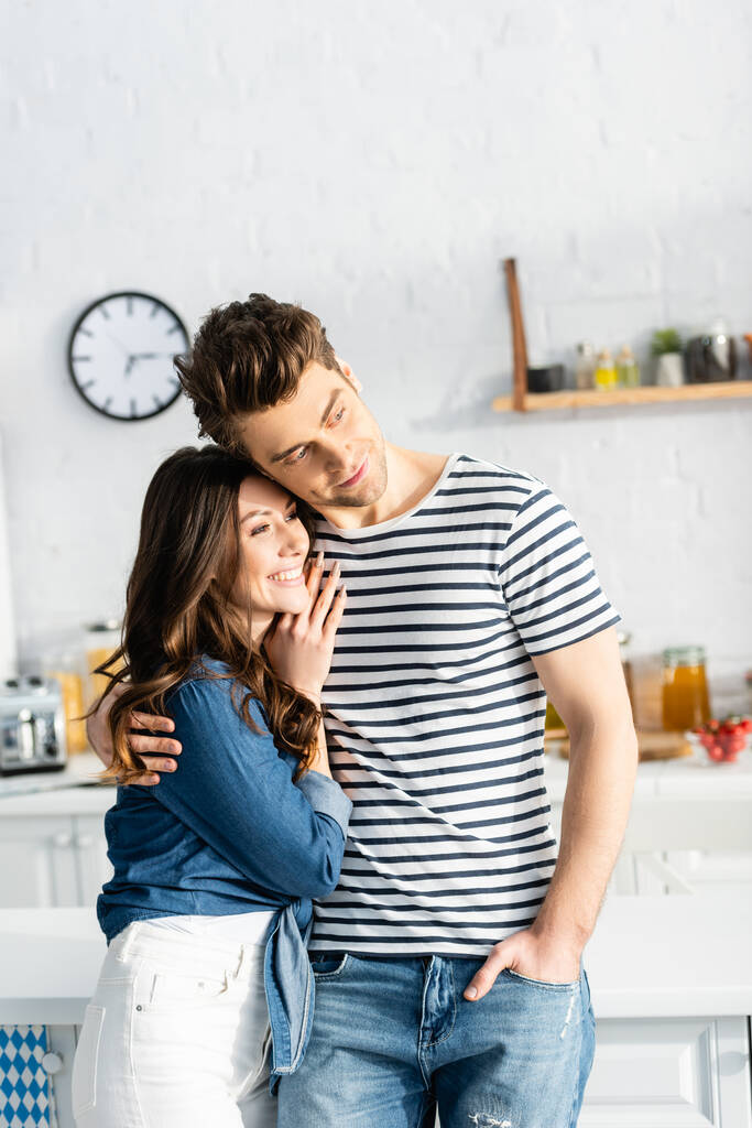 άντρας αγκαλιάζει χαρούμενη φιλενάδα και κοιτάζει αλλού στην κουζίνα  - Φωτογραφία, εικόνα