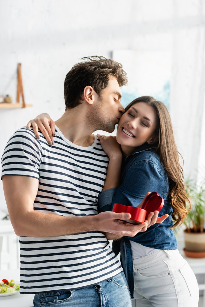 Mann küsst Wange einer glücklichen Freundin, während er herzförmige Geschenkschachtel hält - Foto, Bild
