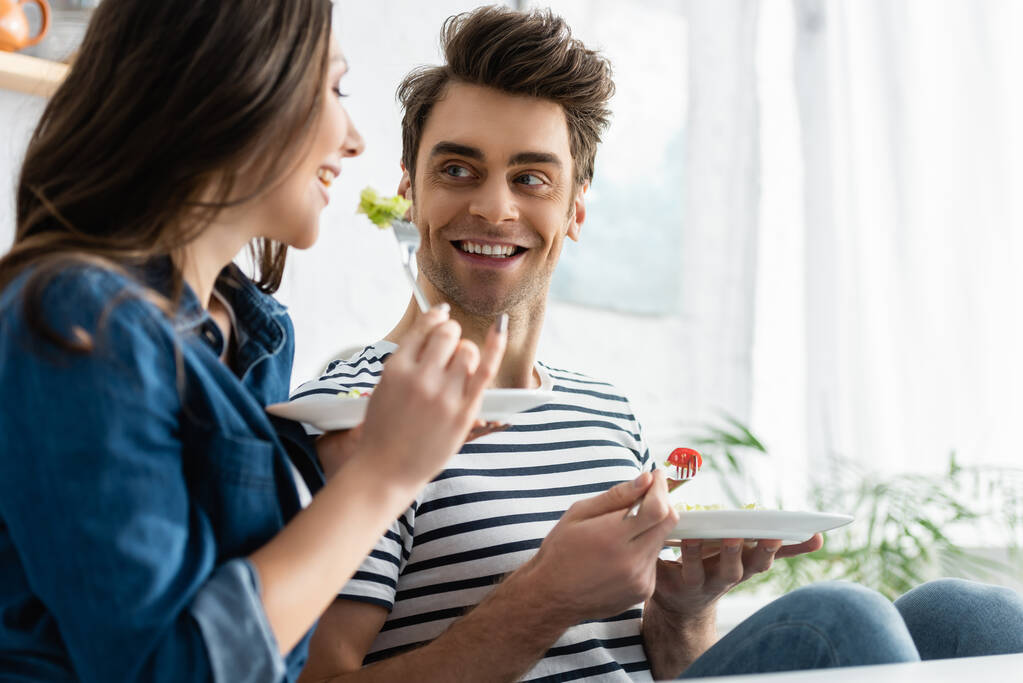homem alegre segurando prato com salada e olhando para a namorada em primeiro plano borrado - Foto, Imagem