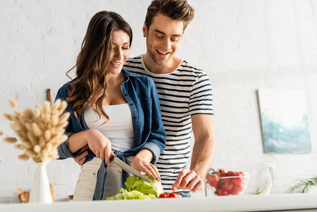 счастливая пара готовит салат на кухне с кеткинами на размытом переднем плане - Фото, изображение