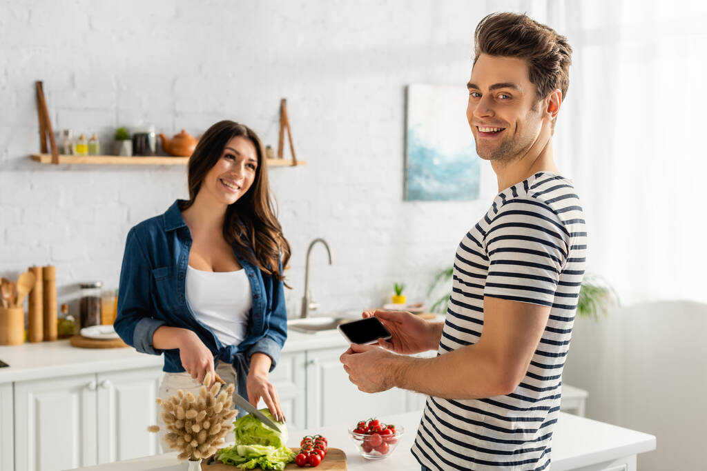 szczęśliwy mężczyzna gospodarstwa smartfon z pusty ekran a kobieta gotowanie w kuchni  - Zdjęcie, obraz