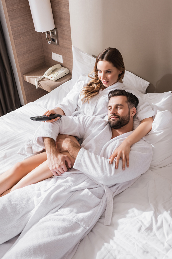 Mulher abraçando namorado em roupão de banho e segurando controlador remoto na cama no hotel  - Foto, Imagem