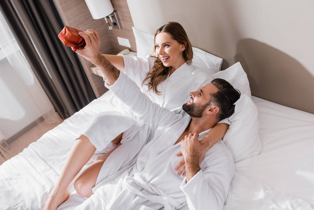 Веселый мужчина в халате держит подарок рядом с девушкой на кровати отеля  - Фото, изображение