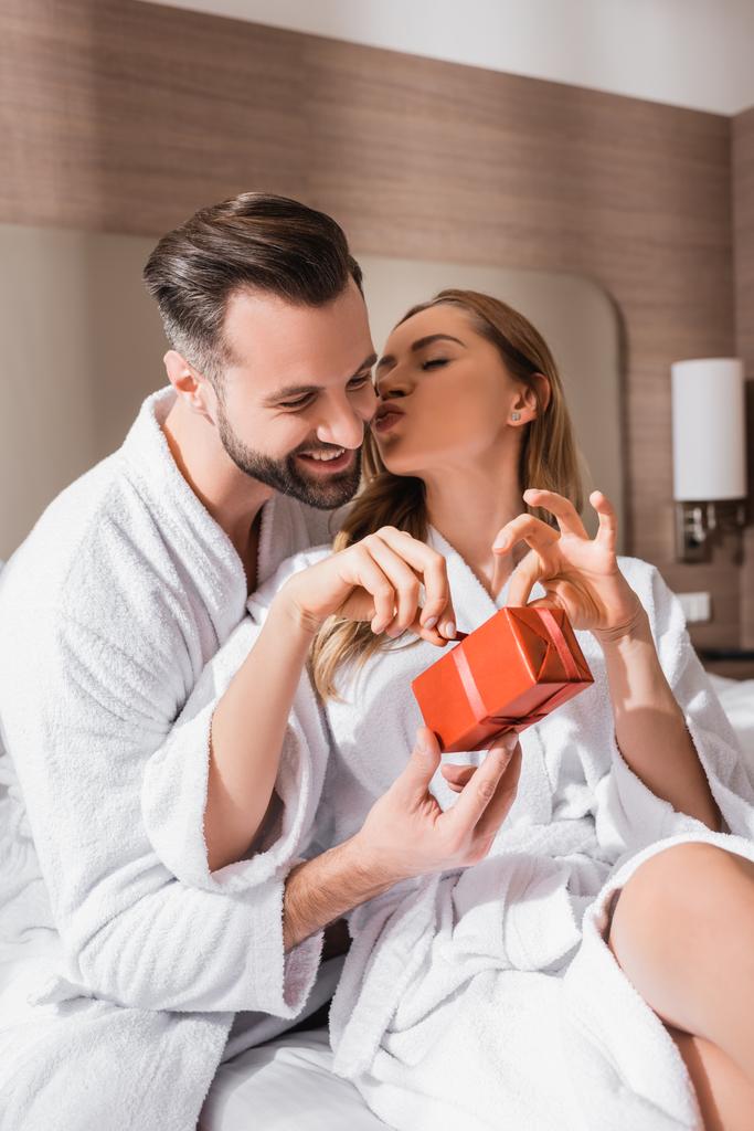 Woman in bathrobe kissing boyfriend while unpacking present on hotel bed  - Photo, Image