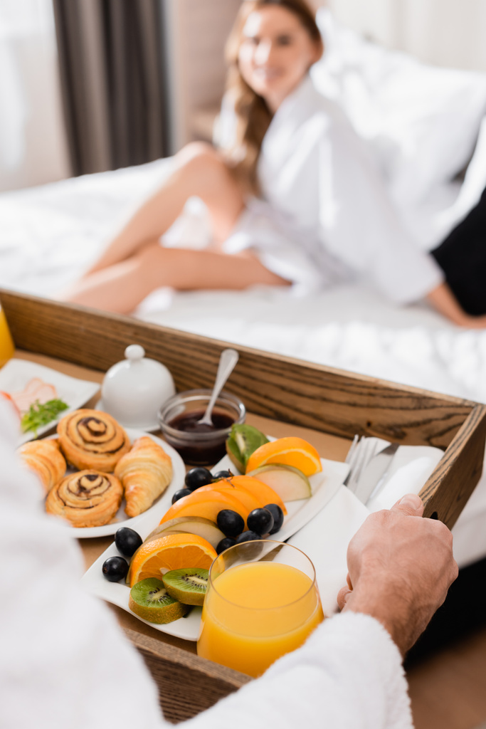 Homme tenant plateau de petit déjeuner avec jus d'orange et fruits près de la petite amie sur le lit de l'hôtel sur fond flou  - Photo, image