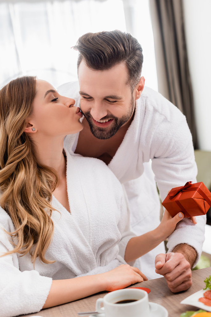 Woman kissing smiling boyfriend and holding present near coffee on blurred foreground in hotel  - Photo, Image