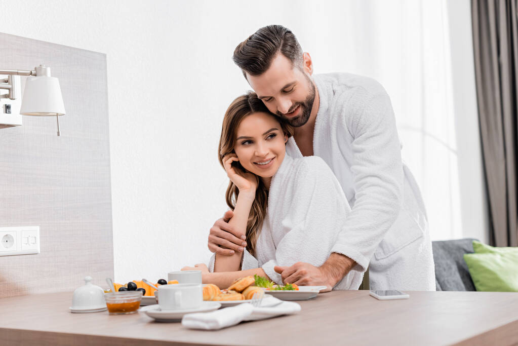 Glimlachende man in badjas knuffelen vriendin in de buurt smakelijk ontbijt in hotelkamer  - Foto, afbeelding
