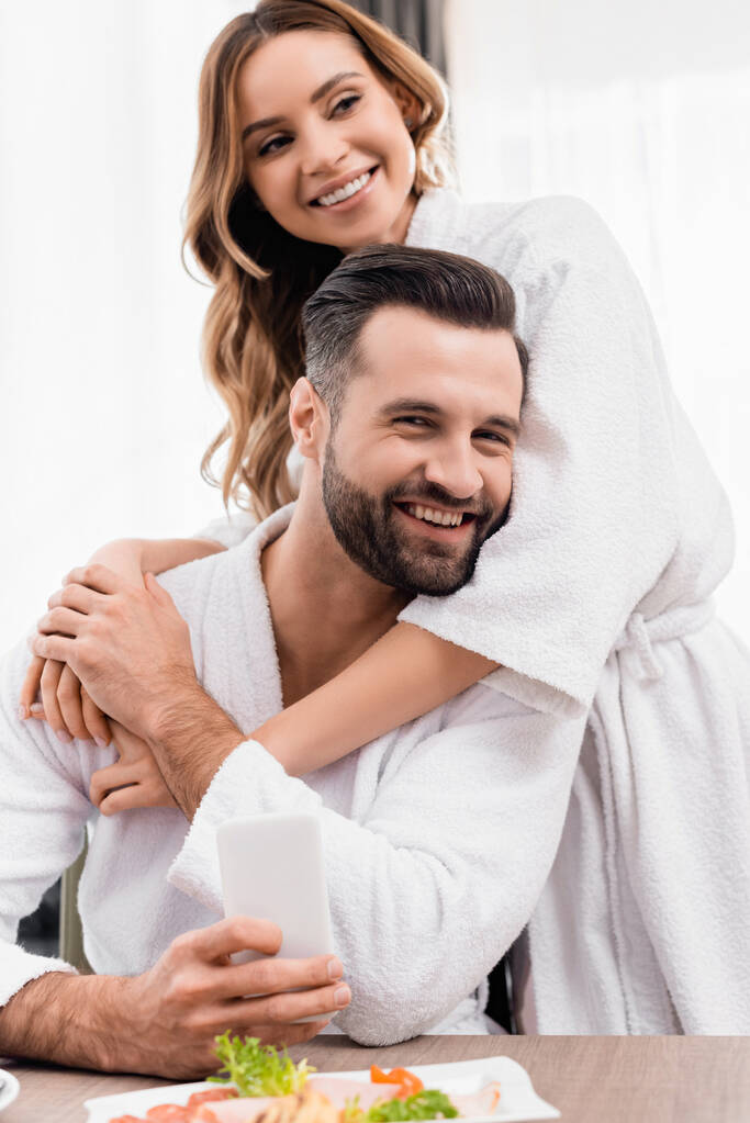 Cheerful woman in bathrobe hugging boyfriend near food on blurred foreground in hotel  - Photo, Image