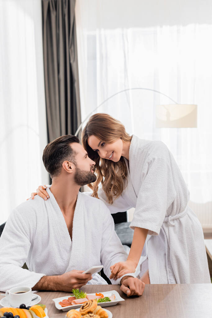 Mujer sonriente abrazando novio en albornoz con teléfono inteligente cerca del desayuno y café en primer plano borroso en la habitación del hotel  - Foto, imagen