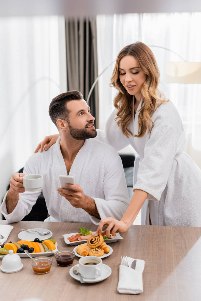 Nainen ottaa leivonnaisia lähellä poikaystävä kuppi kahvia ja matkapuhelin hotellihuoneessa  - Valokuva, kuva