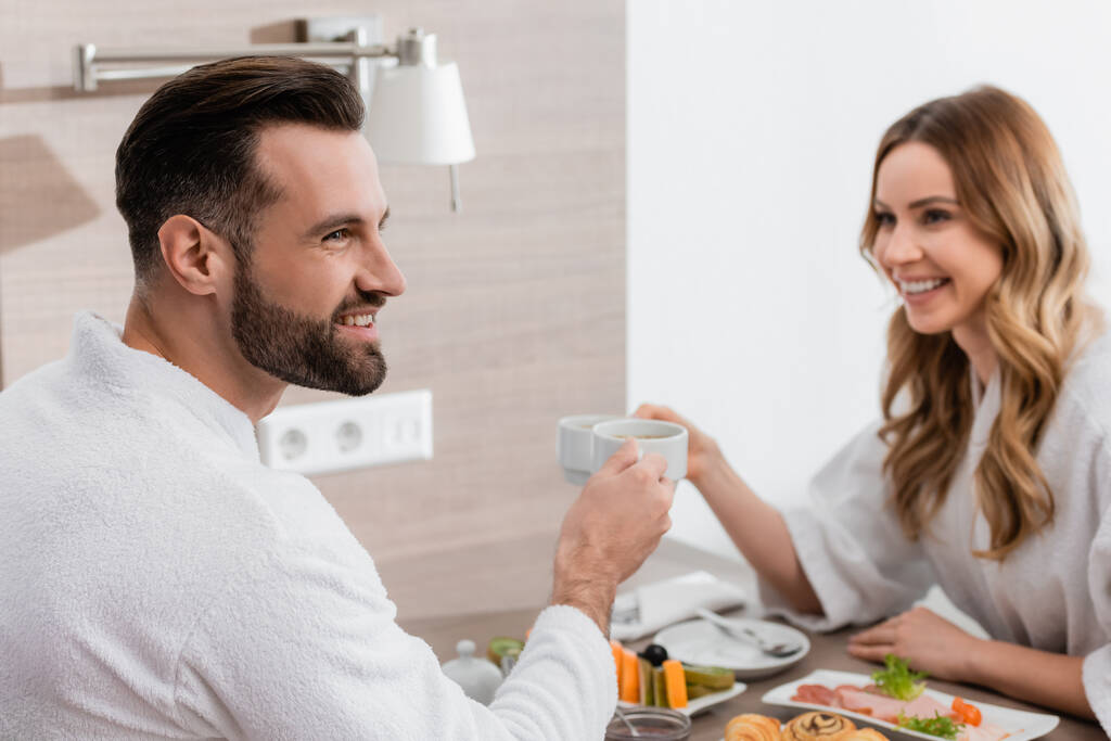 Улыбающийся мужчина в халате держит чашку возле завтрака и подружку на размытом фоне в отеле  - Фото, изображение