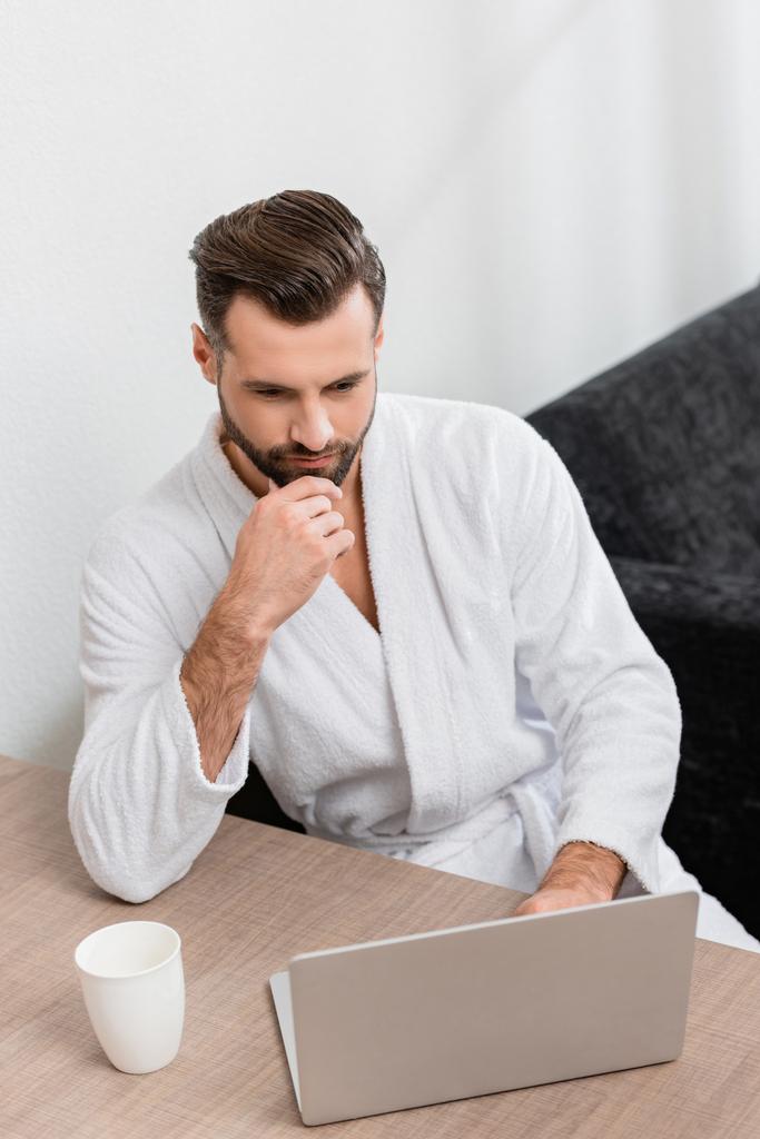 Freelancer in bathrobe looking at laptop near cup in hotel  - Photo, Image