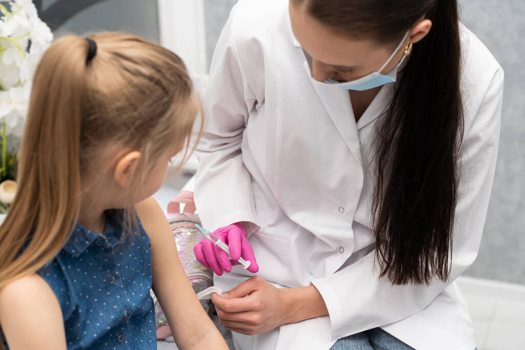 Una enfermera se está preparando para inyectar a una niña con una vacuna previamente preparada. El joven aprendiz intenta hablar con el niño para que no tenga miedo de la inyección. - Foto, Imagen