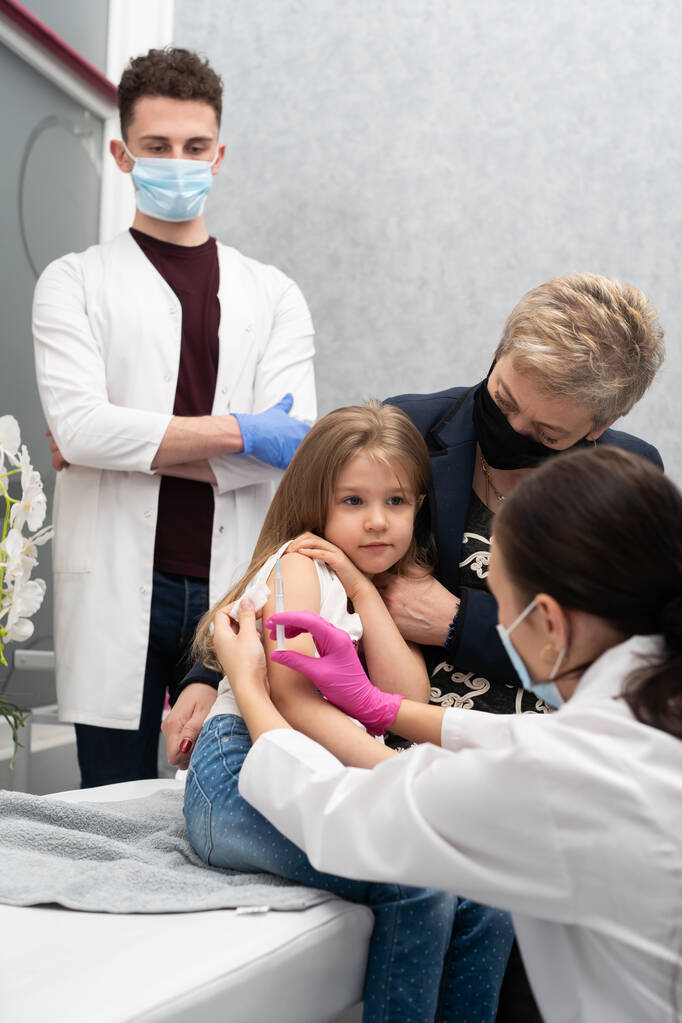 La abuela saluda a su nieta durante la vacunación. Una enfermera joven apuñala una aguja en el brazo de las chicas. Un aprendiz en el último año de medicina observa toda la situación en el consultorio médico - Foto, Imagen