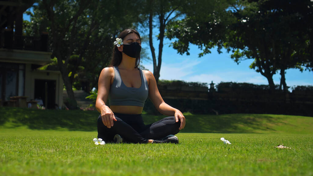 Joven morena europea practica yoga sola en la naturaleza sentada sobre hierba verde en pose de loto usando una máscara protectora negra en su cara - Foto, imagen